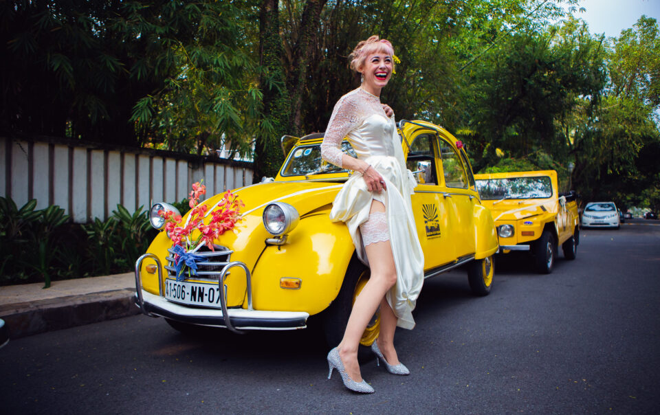 Bride laughing and showing her garter in front of two yellow cars just before heading to her wedding.