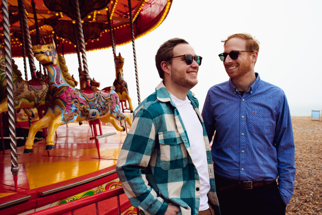 LGBTQ+ couple smiling during their engagement shoot next to the horse carousel on Brighton beach