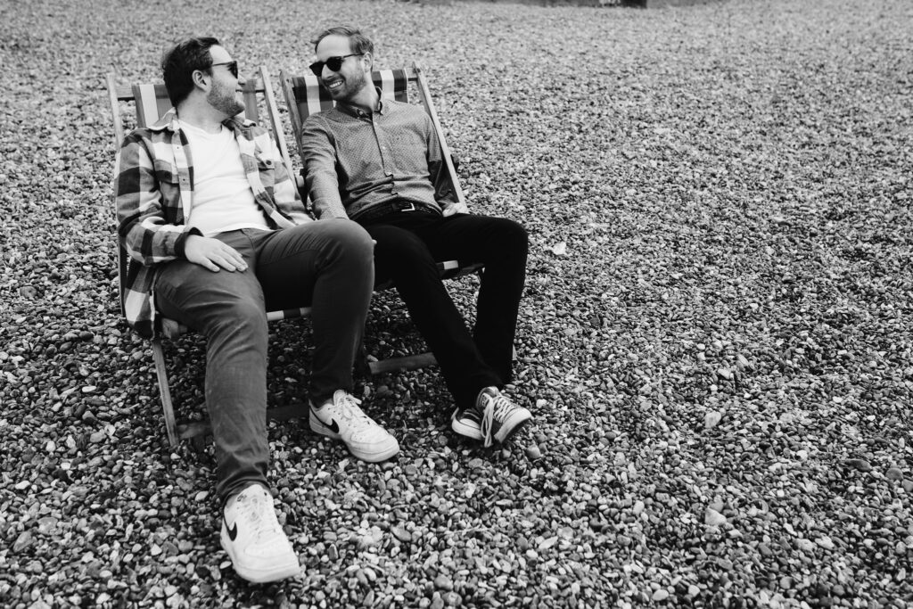 LGBTQ+ couple looking at each other and holding hands on deck chairs at Brighton beach