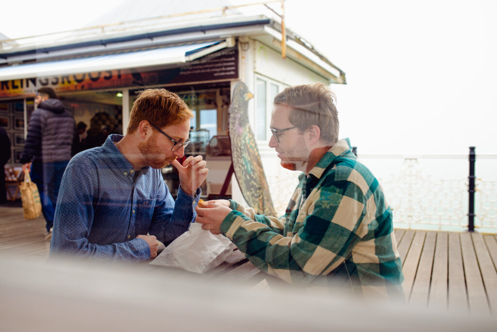 LGBTQ+ couple sitting at a picnic table laughing and eating doughnuts on Brighton pier