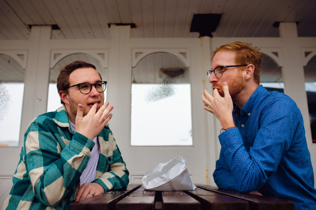 LGBTQ+ couple sitting at a picnic table licking sugar of their fingertips during their engagement session