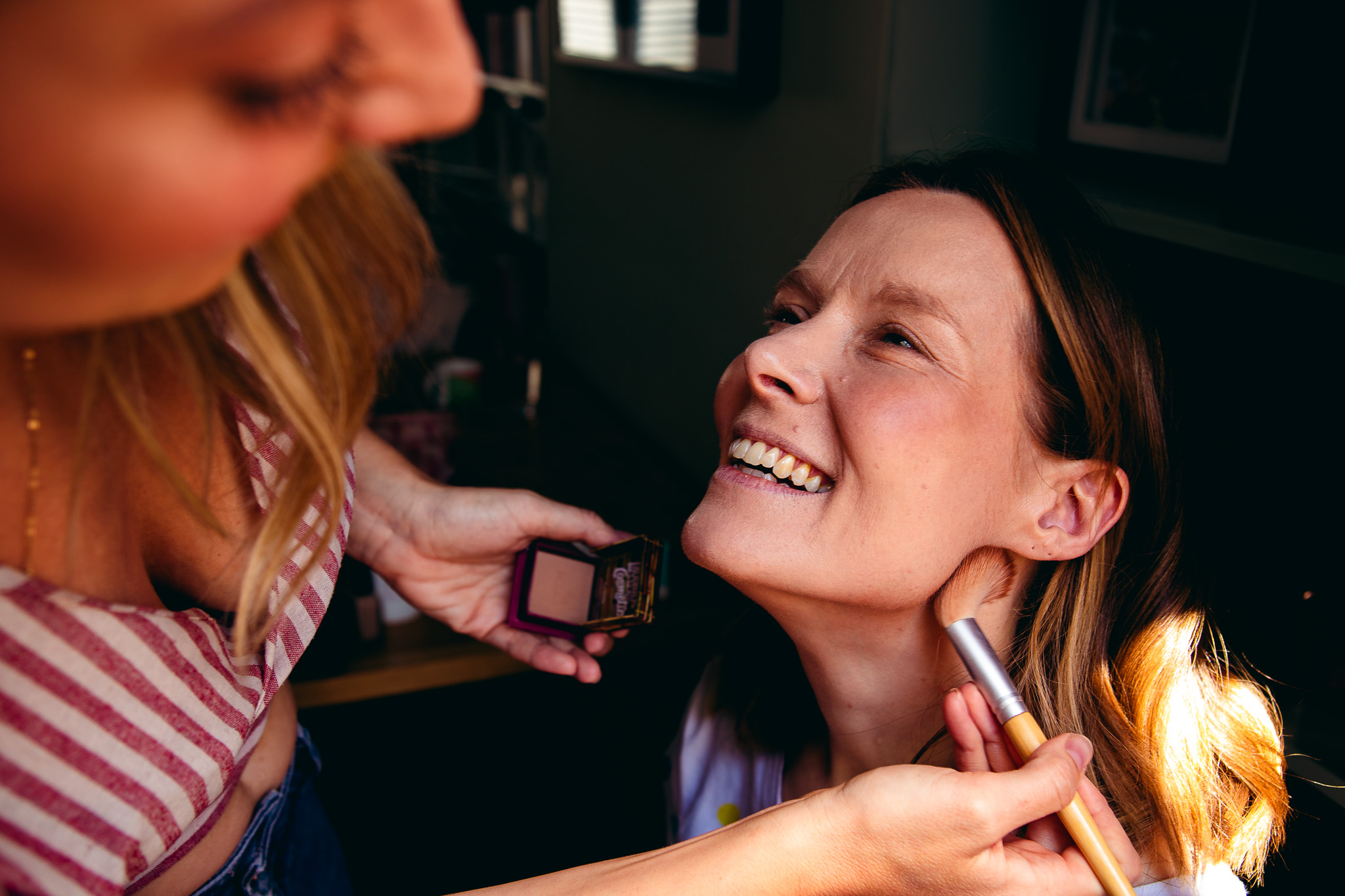 Gemma looks up and smiles at her friend who's applying her bridal makeup in preparation for her wedding day