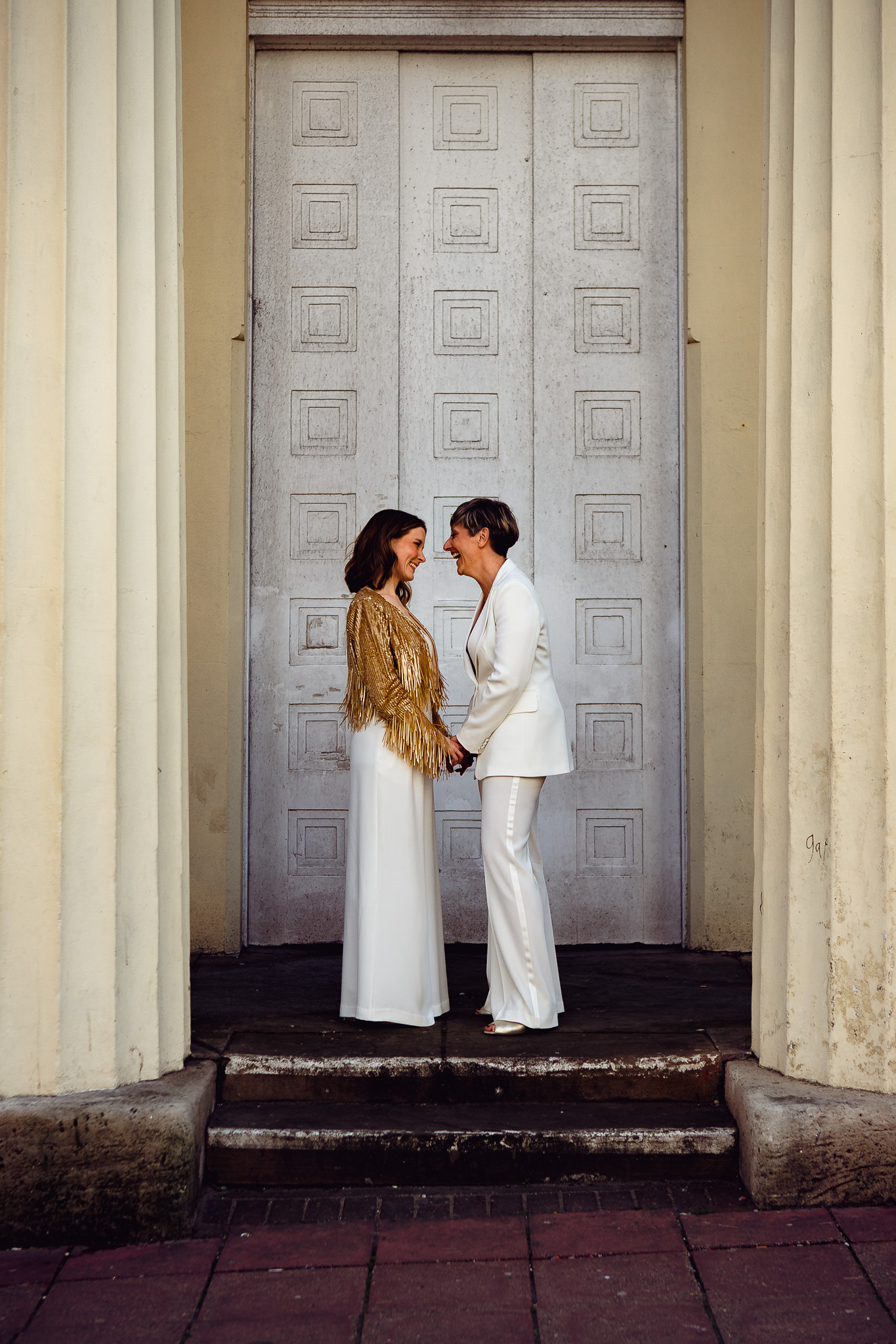 Gemma and Natalie hold hands as they face each other and smile outside Brighton Town Hall during their couple portraits.