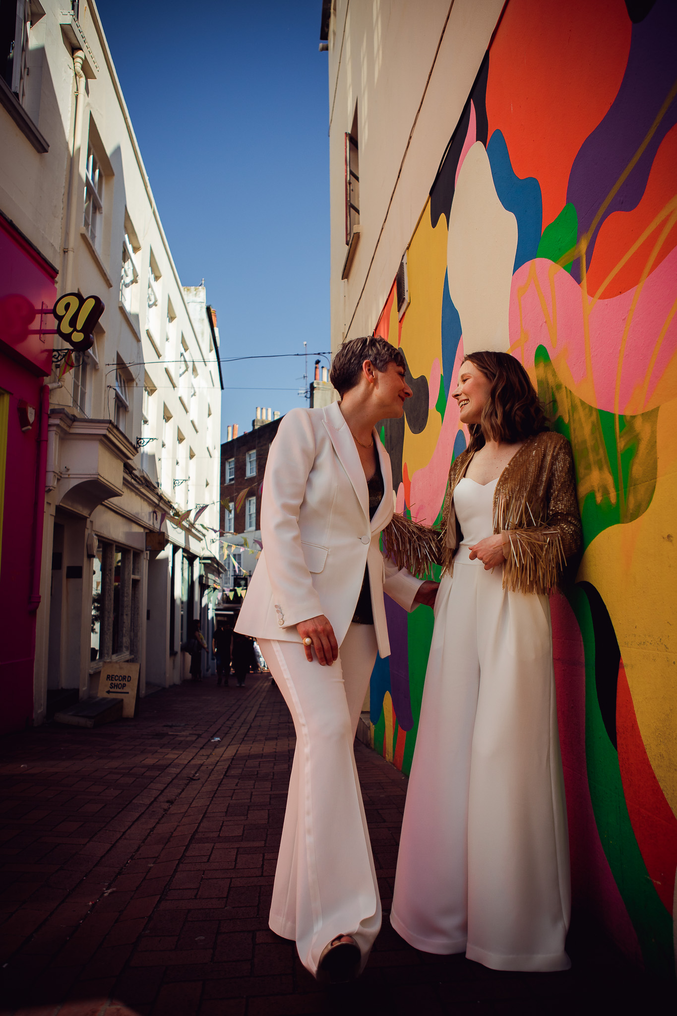 Gemma and Natalie stand together smiling next to a colourful mural in the Brighton lanes during their couple portraits.