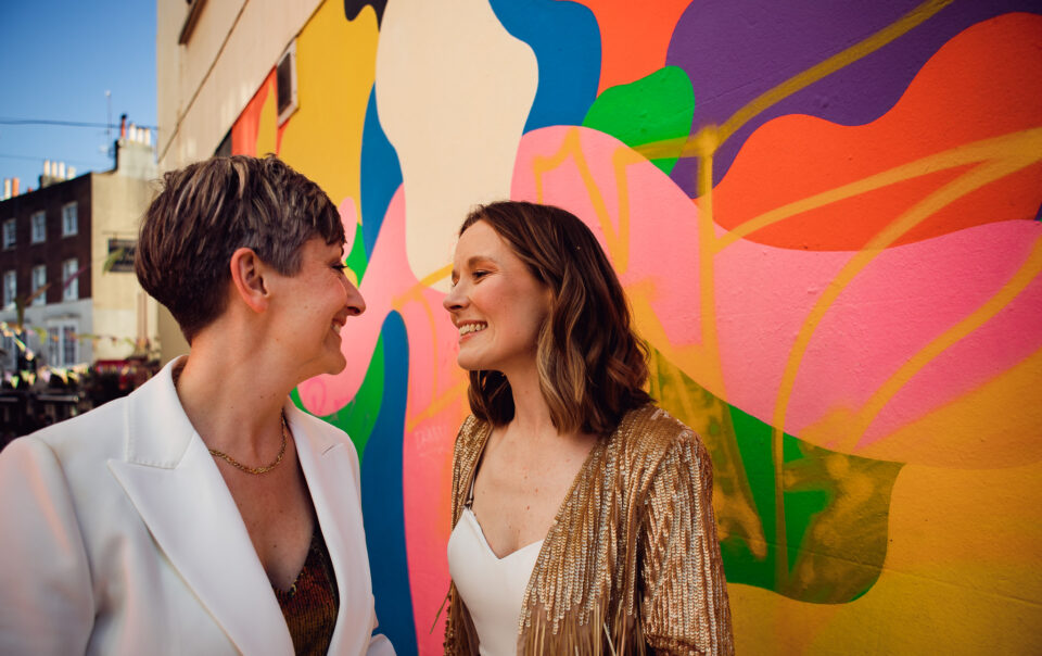 Gemma and Natalie smile at each other next to a colourful mural in the Brighton lanes during their couple portraits.