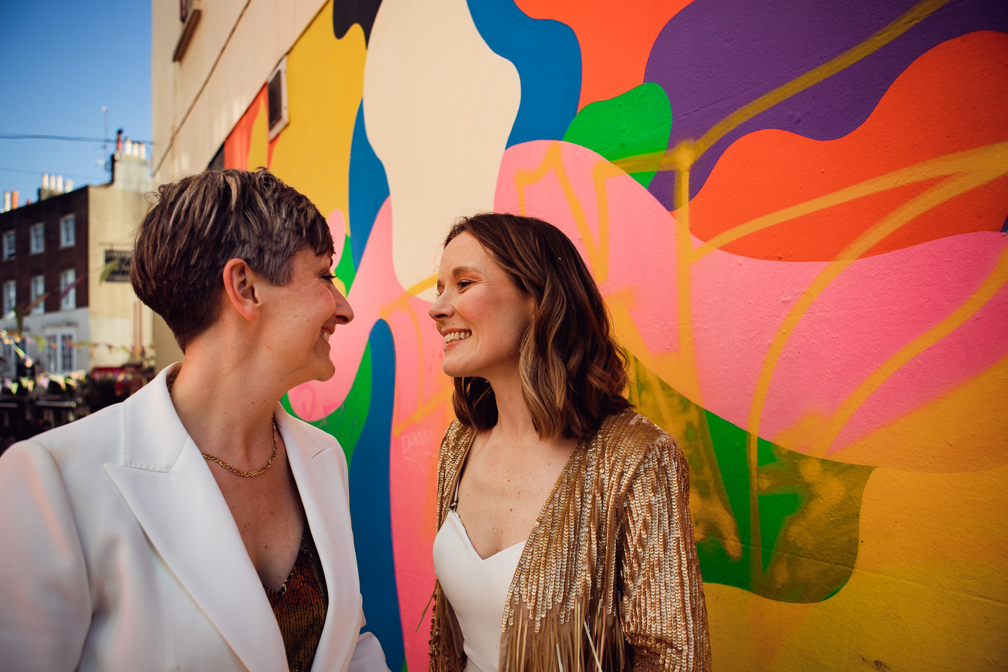 Gemma and Natalie smile at each other next to a colourful mural in the Brighton lanes during their couple portraits.