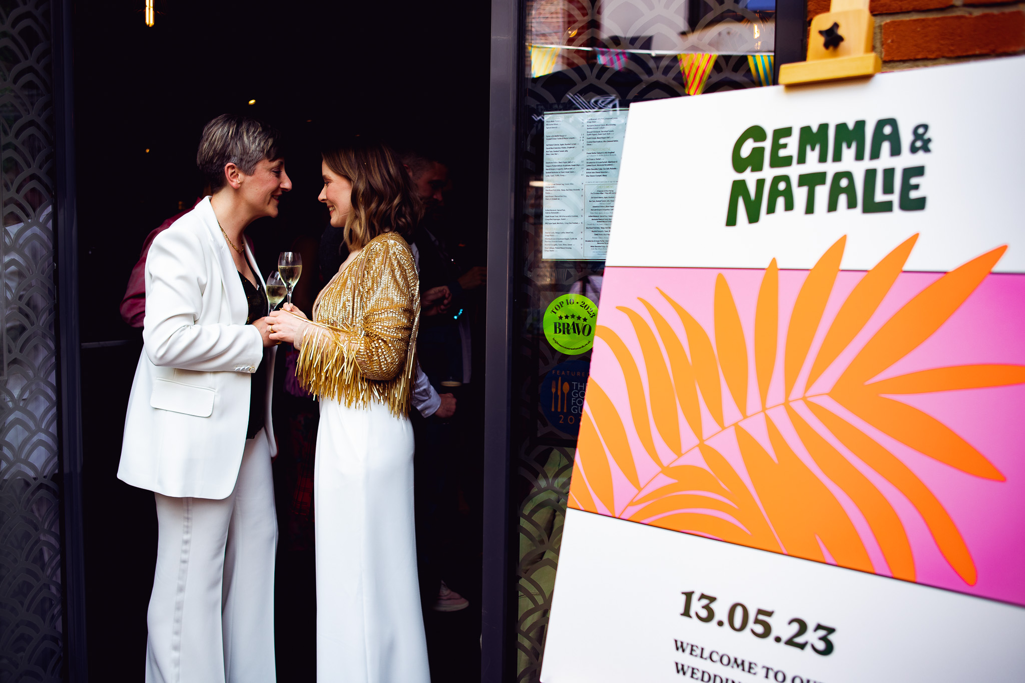 Gemma and Natalie stand looking at each other in the doorway of the Flint House next to a colourful wedding welcome poster.