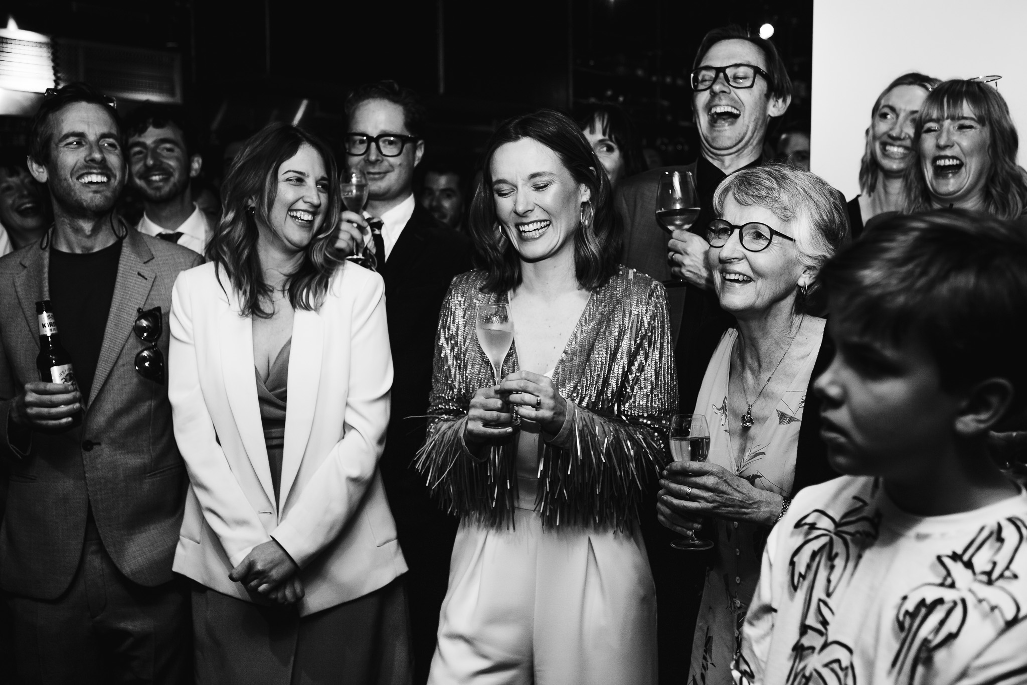 Gemma smiles surrounded by her laughing wedding guests during the wedding speeches.