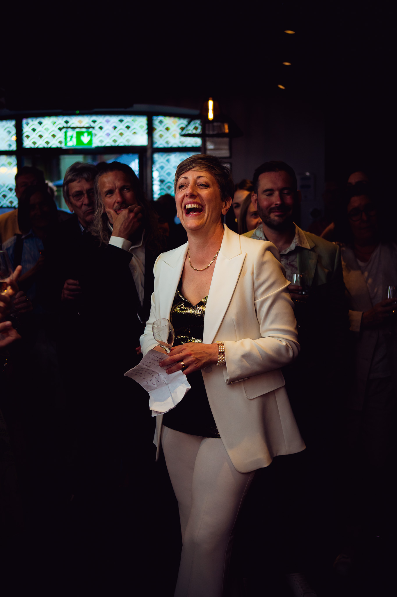 Natalie laughs as she stands amongst guests and listens to a wedding speech.