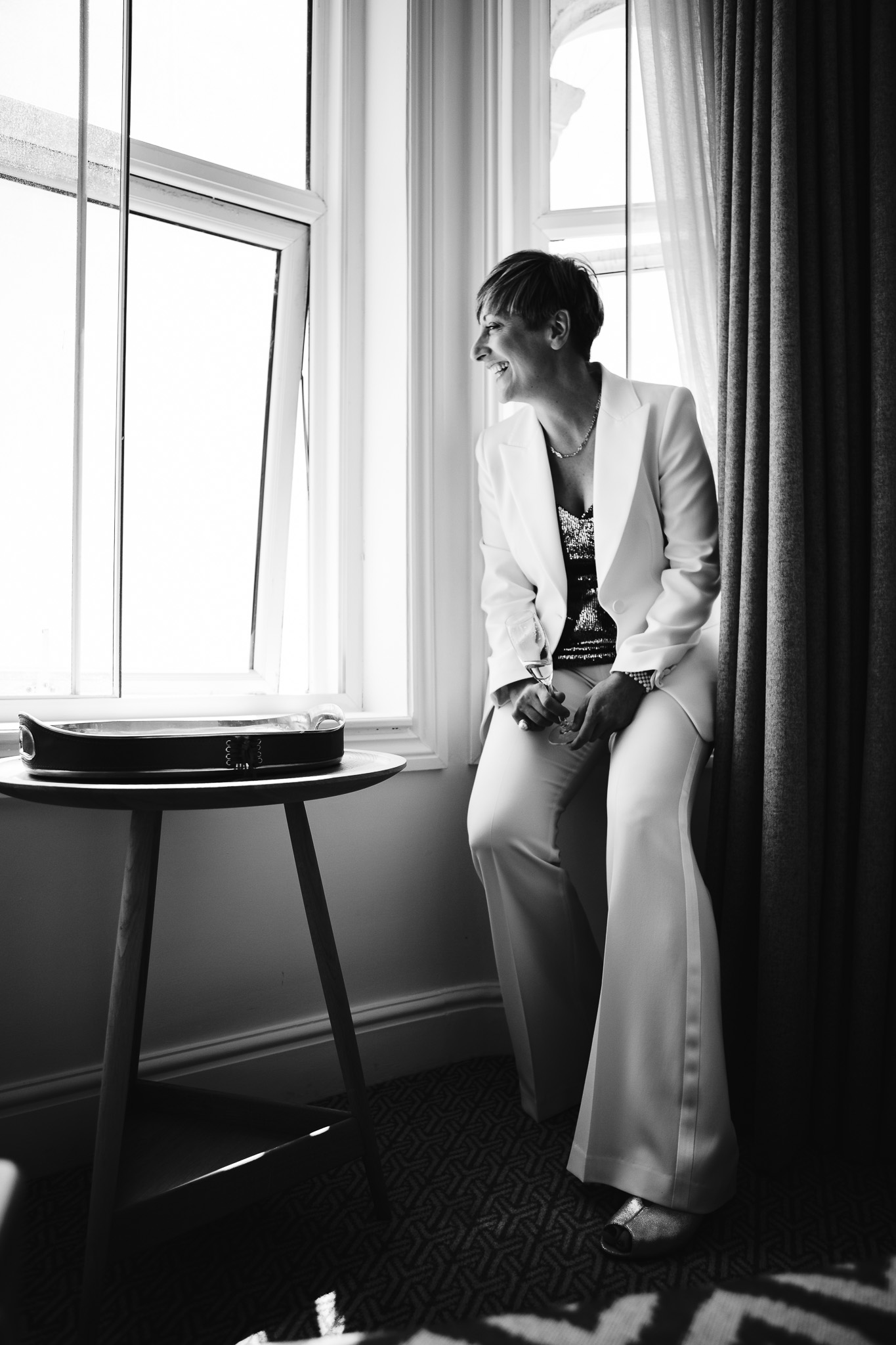 Natalie sits smiling by the window in The Harbour Hotel, Brighton, holding a champagne flute in preparation for her wedding.